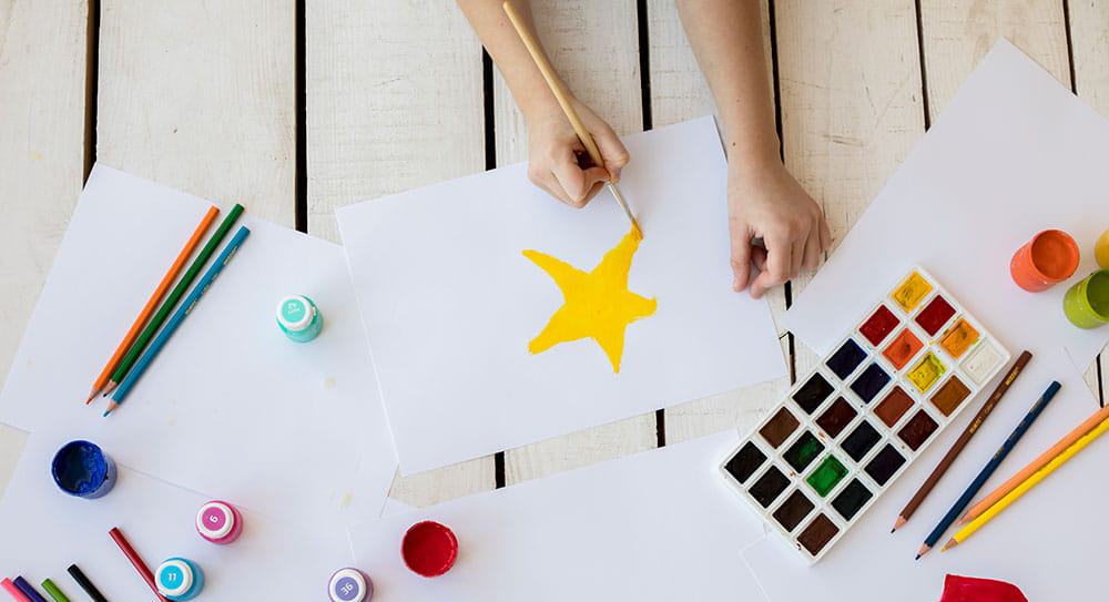 overhead-view-girl-painting-yellow-star-with-paintbrush-white-paperCropped