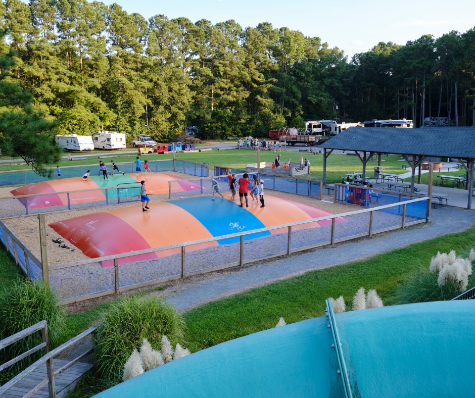 Children playing on large inflatable jumping pillows in a fenced play area at a family-friendly campground, surrounded by greenery and RVs.