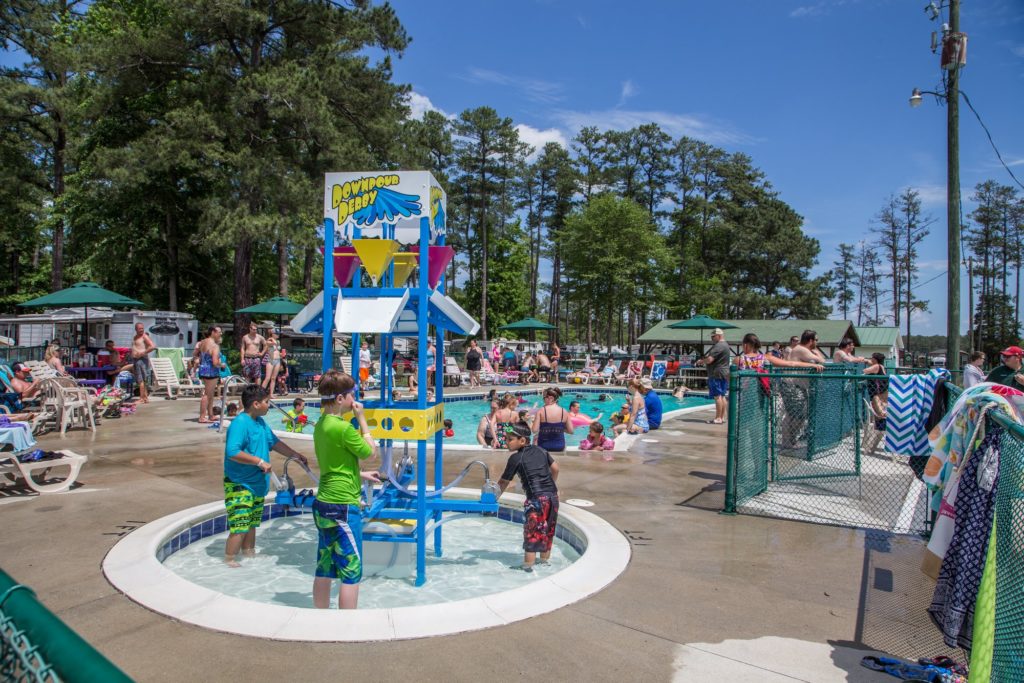 Children playing in a shallow splash area with interactive water features at a busy campground pool, surrounded by families enjoying the summer sun.