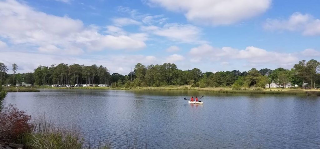 Two people kayaking on a calm lake surrounded by greenery, with a campground visible in the background under a partly cloudy sky.