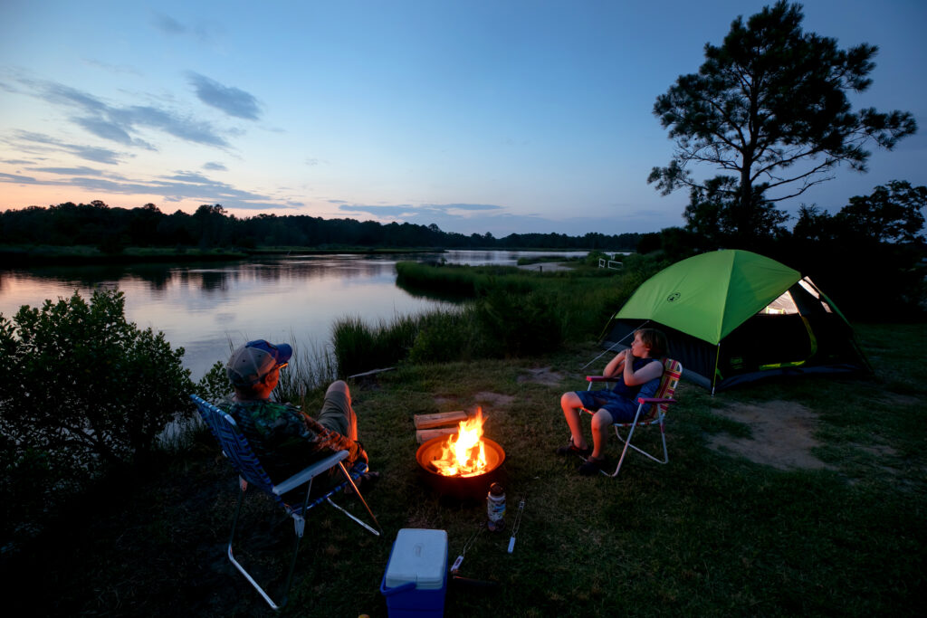 Campers relaxing by a campfire near a green tent with a lakeside view at Camp Cardinal.