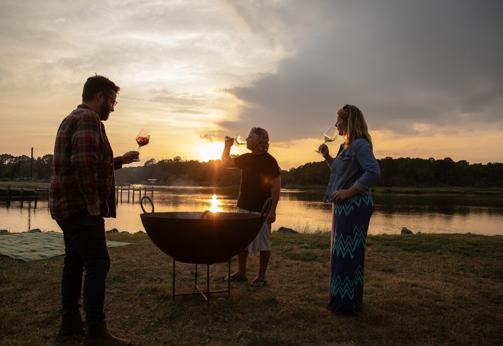 Group enjoying Virginia wine at a lakeside fire pit during sunset at Camp Cardinal.