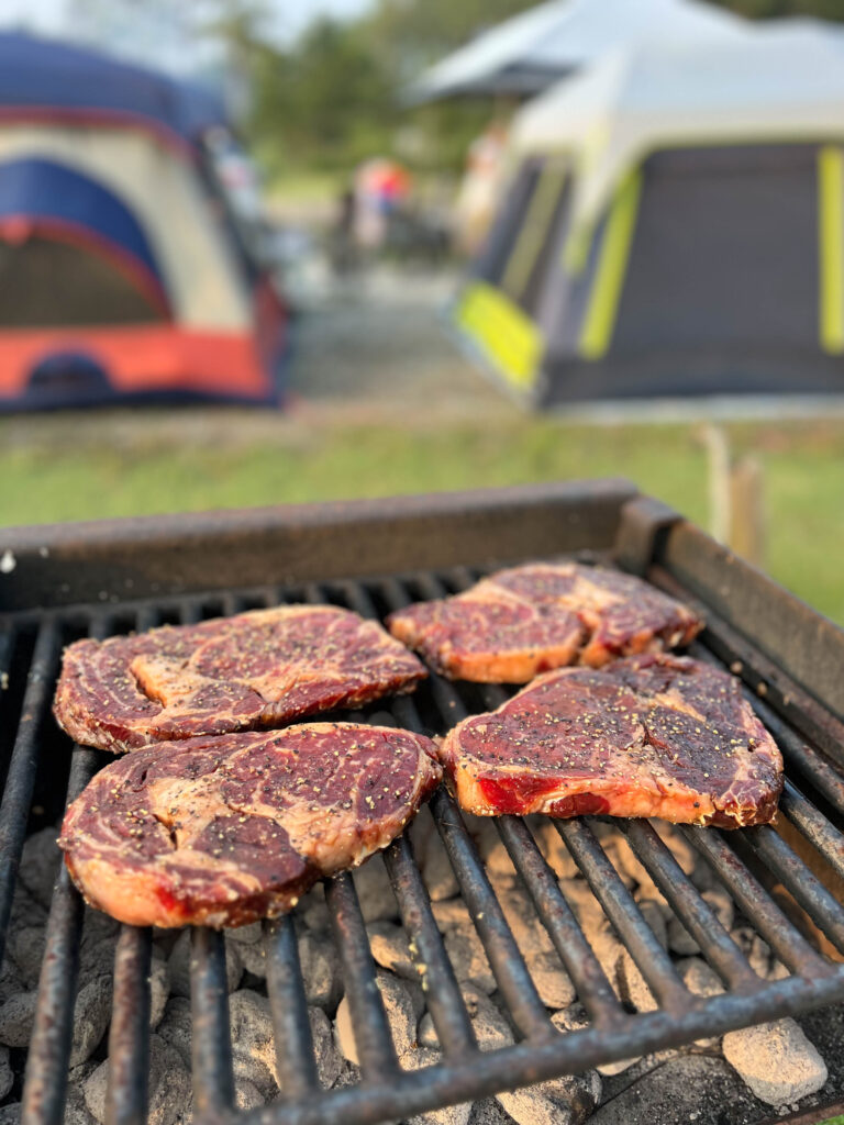 Close-up of seasoned steaks on a grill with tents in the background