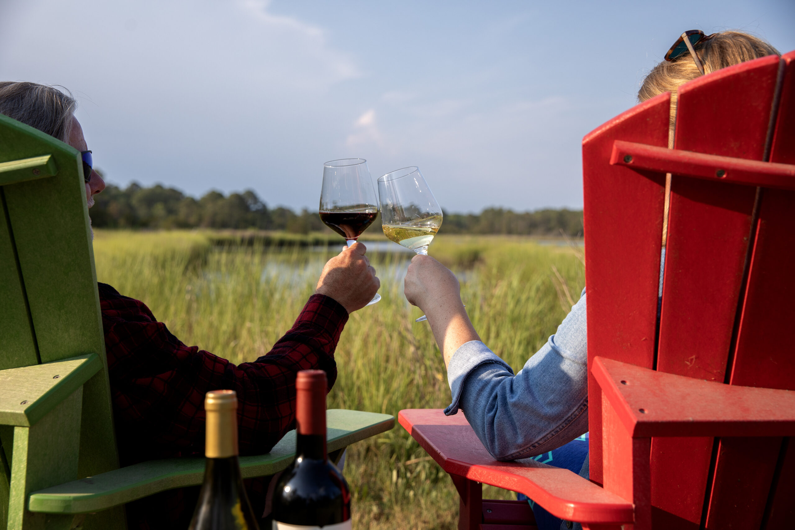 Two people clinking wine glasses in Adirondack chairs overlooking the marsh