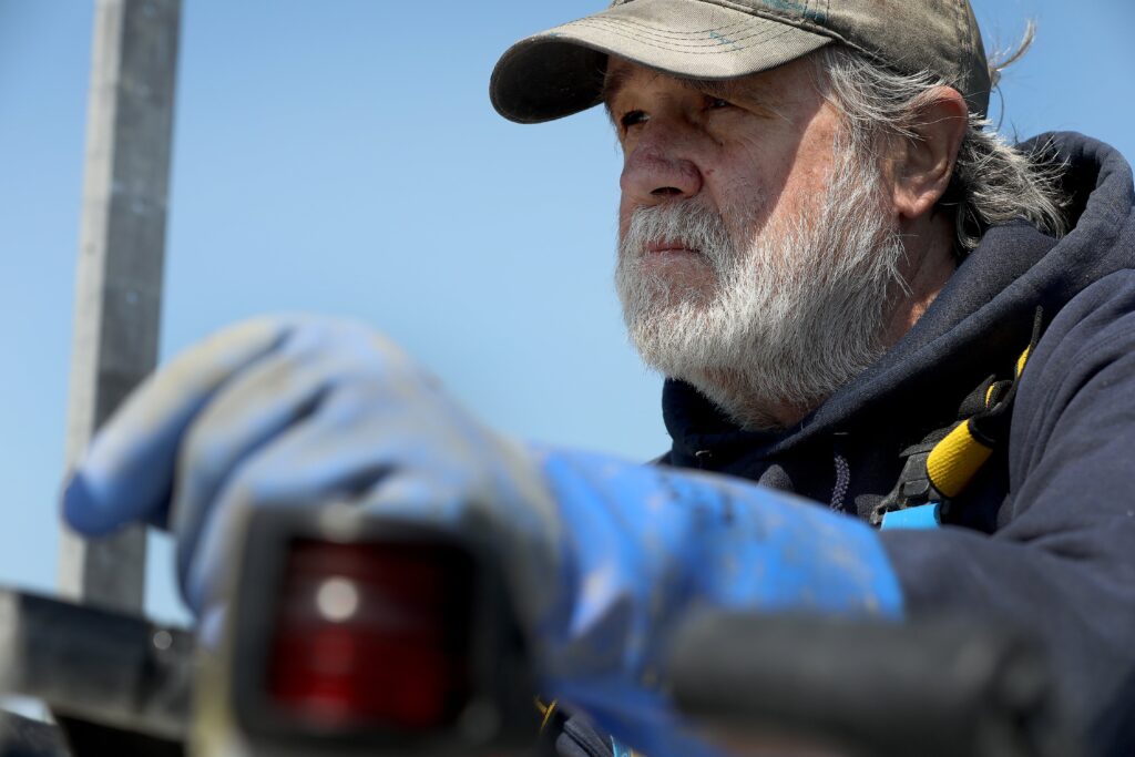 Close-up of a bearded oyster harvester in a hat and gloves working on the water.